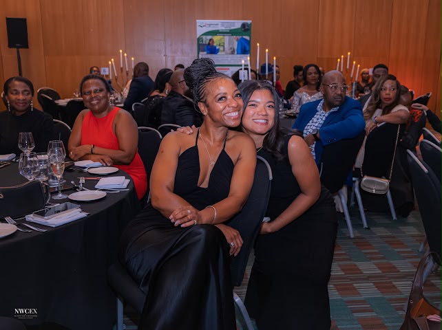 Two women smiling together at the dinner table, with candlelit tables behind them.